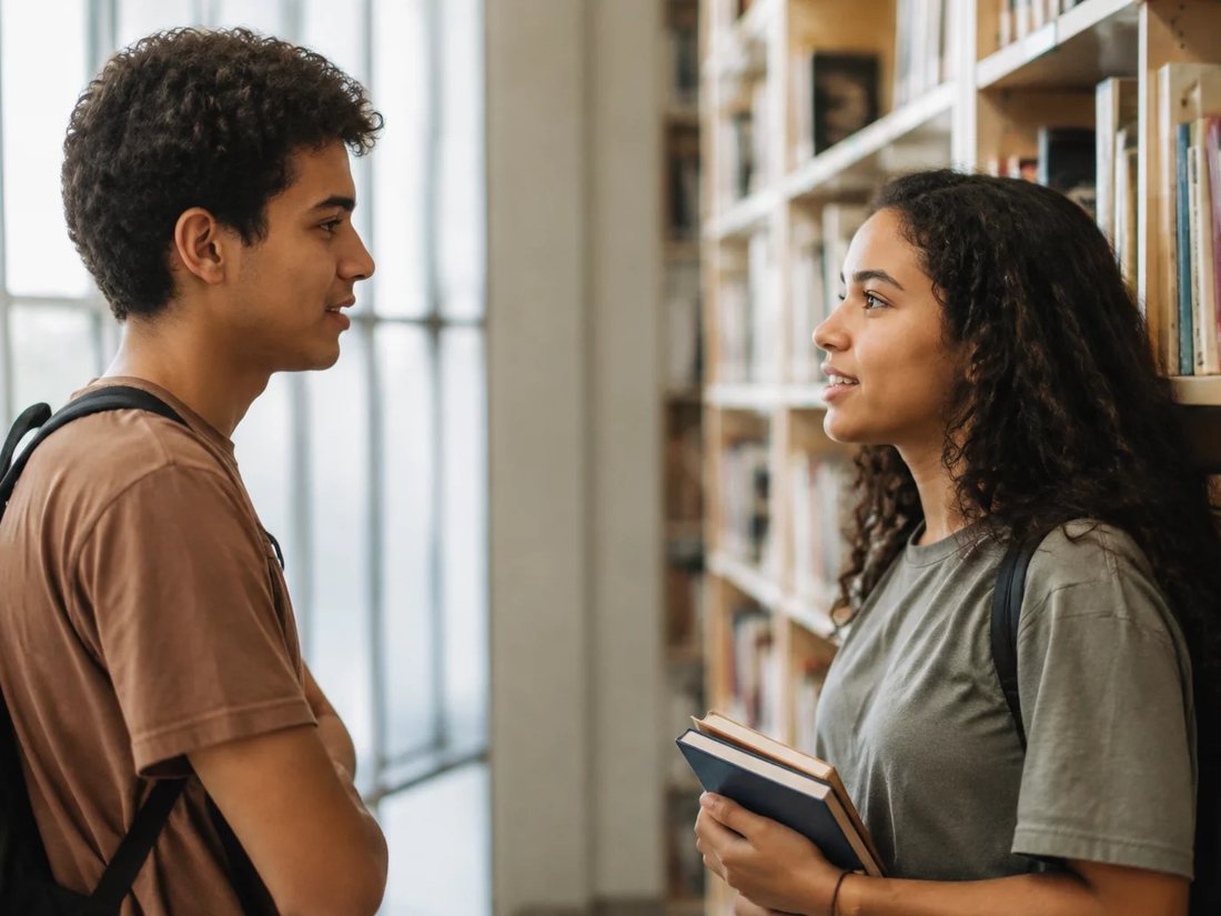 Conversa em pé entre as estantes da escola, com folhas de anotação na mão de quem discute